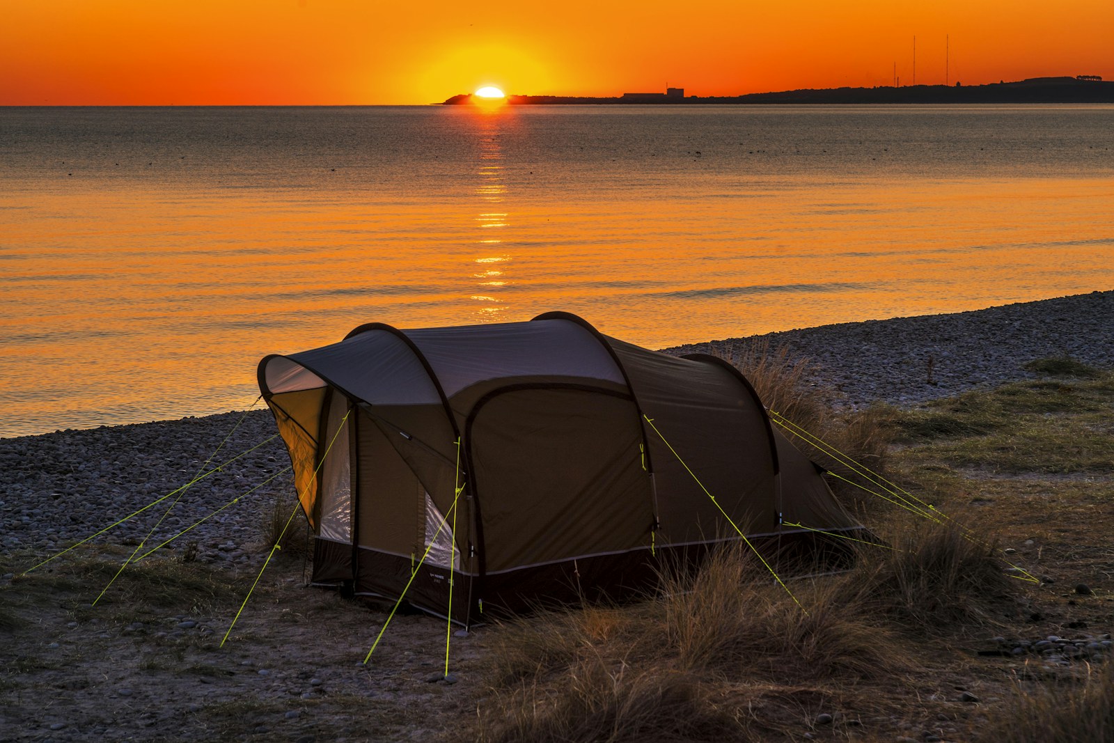 A dark camping tent beside the sea as the sun sets over a pebble beach