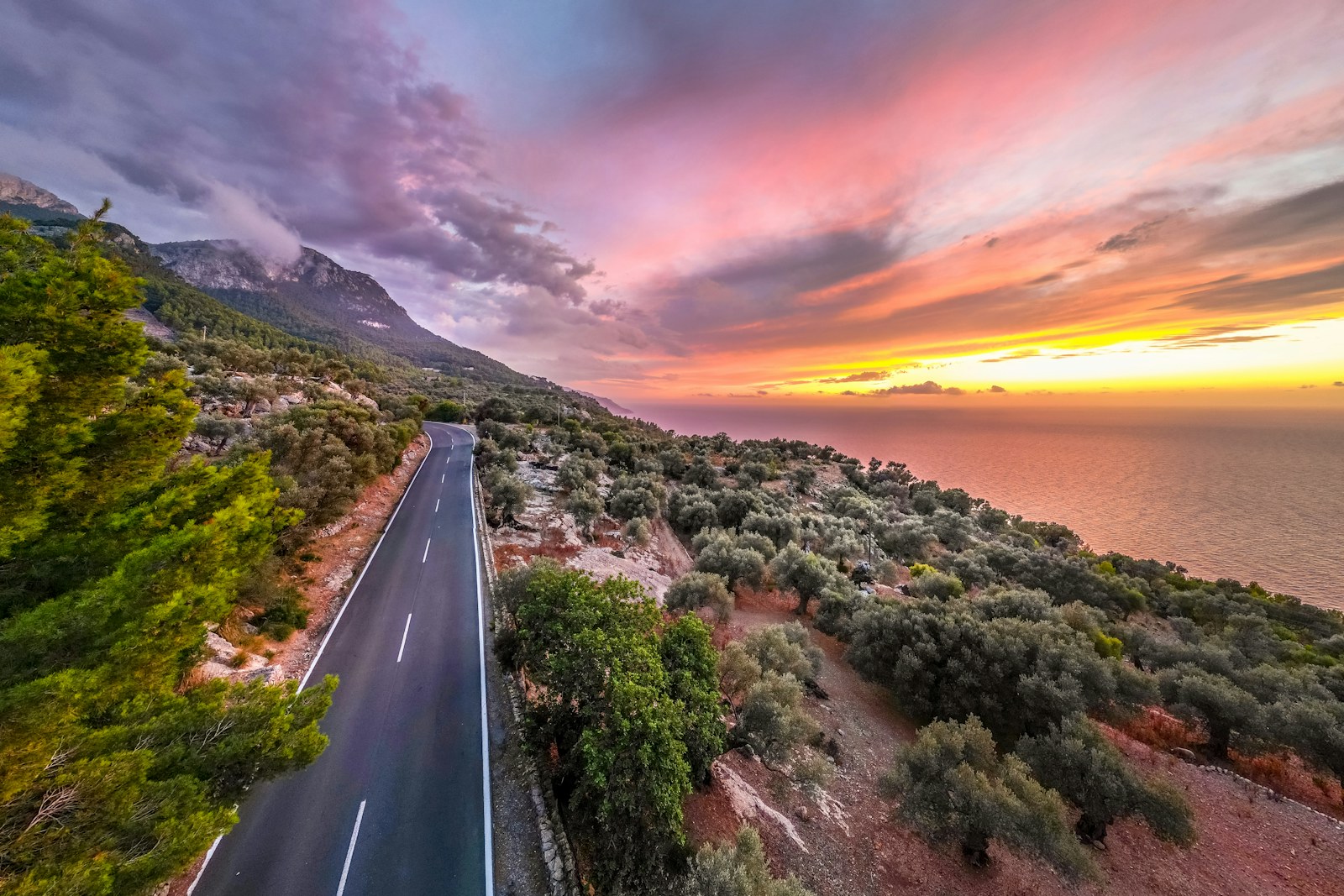 A winding coastal road beside the sea beneath a pink and golden sunset sky