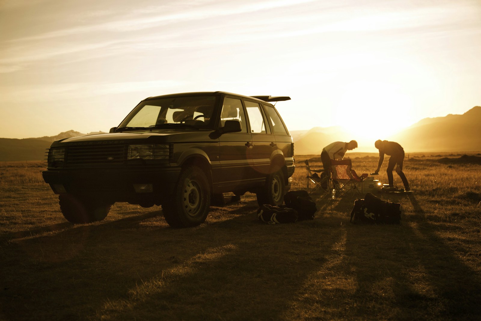 Two travellers arrange gear beside a parked vehicle in warm sunset light in Iceland